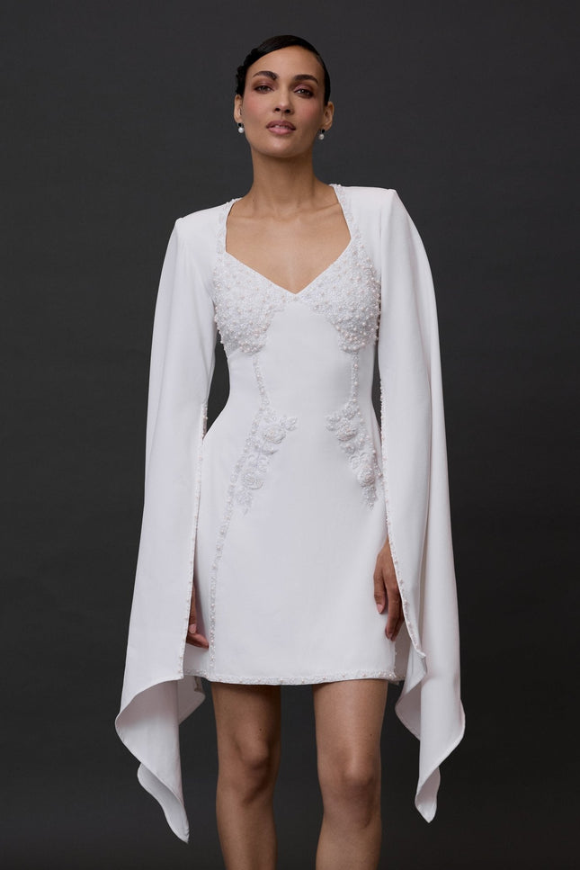 A woman modeling a white mini dress with long sleeves and a V-neckline, featuring a flowing skirt.