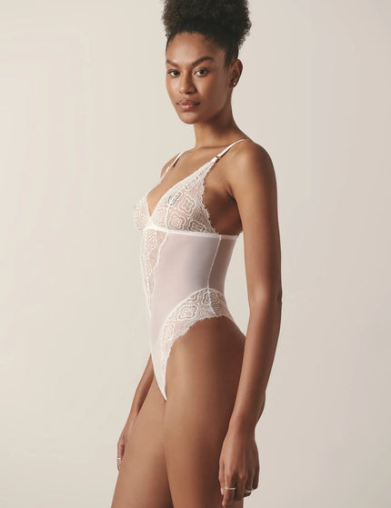 Side view of a model wearing a white mesh and lace lingerie bodysuit, posing in a studio