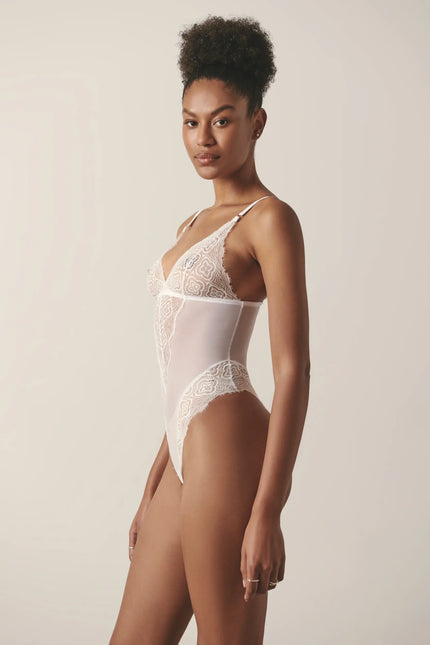 Side view of a model wearing a white mesh and lace lingerie bodysuit, posing in a studio